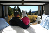 Inside their RV rental, a father and child gaze at a stunning mountain, forest, and river view. Perfect road trip stop.