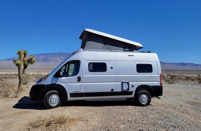 Silver pop-top camper van (RV rental) parked on a dirt road in a sunny desert, with a Joshua tree and distant mountains. Perfect for adventure.