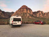 A white RV rental van and a red car are parked on a gravel lot, with majestic red rock mountains visible under a sunset sky.