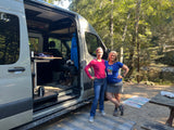 Two friends beside their custom rv rental van, open to show interior. By a river in a wooded campsite.