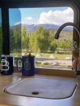 Serene RV rental kitchen view with mugs and sink. Outside, mountains, forest, and sky visible through the window screen.