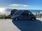 Enjoy an RV rental: a dark grey camper van parked by a sunny beach and ocean view. Ideal for road trip adventures!