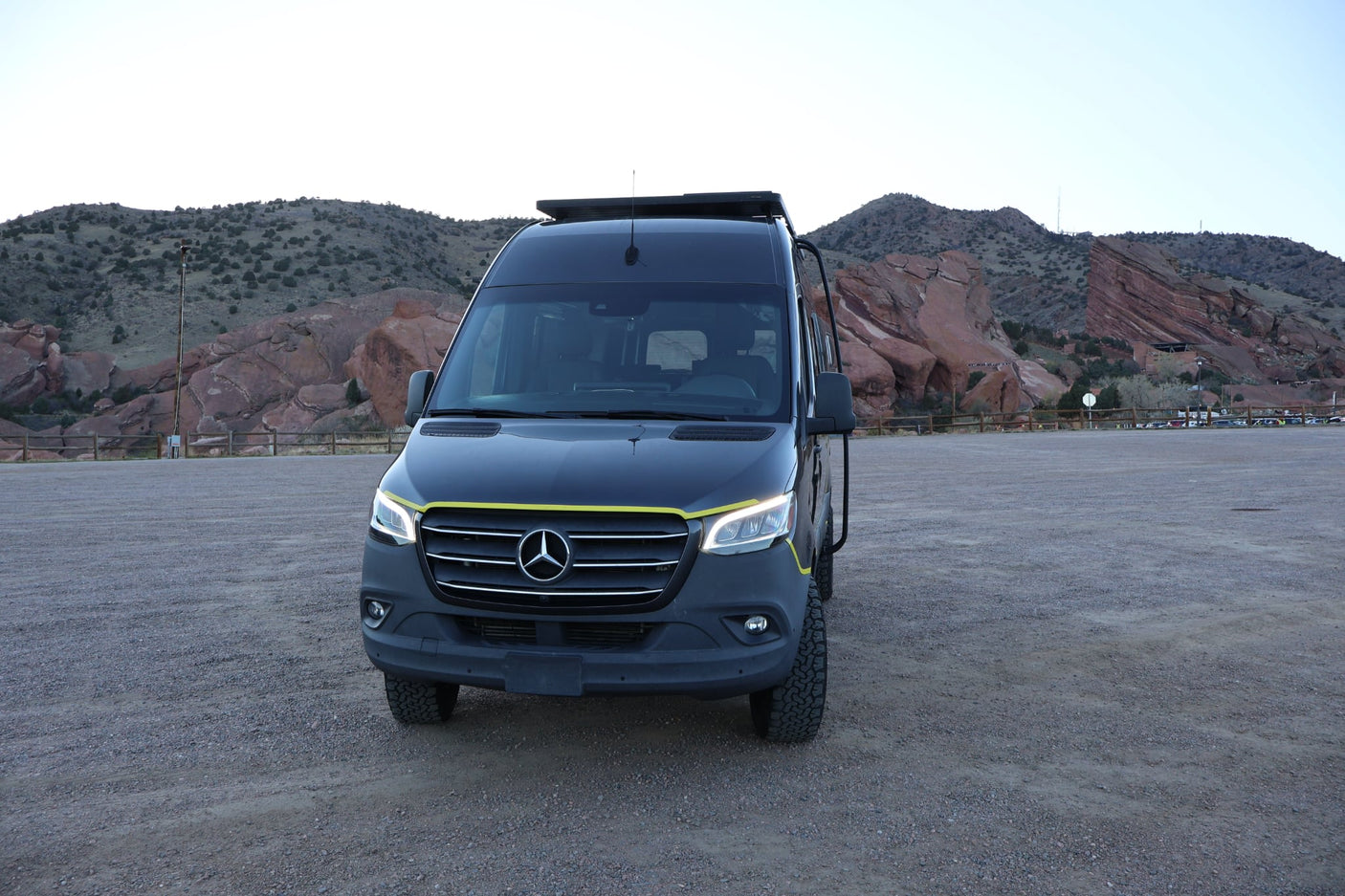 Adventure-ready Mercedes Sprinter RV rental, with a yellow stripe, parked on gravel before scenic red rock formations.