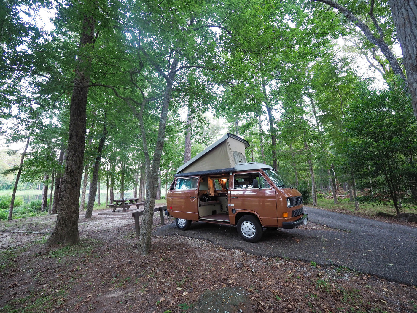 Experience camping! Brown RV rental (VW camper van) parked in a lush forest campsite by a lake. Interior light on.
