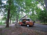 Experience camping! Brown RV rental (VW camper van) parked in a lush forest campsite by a lake. Interior light on.