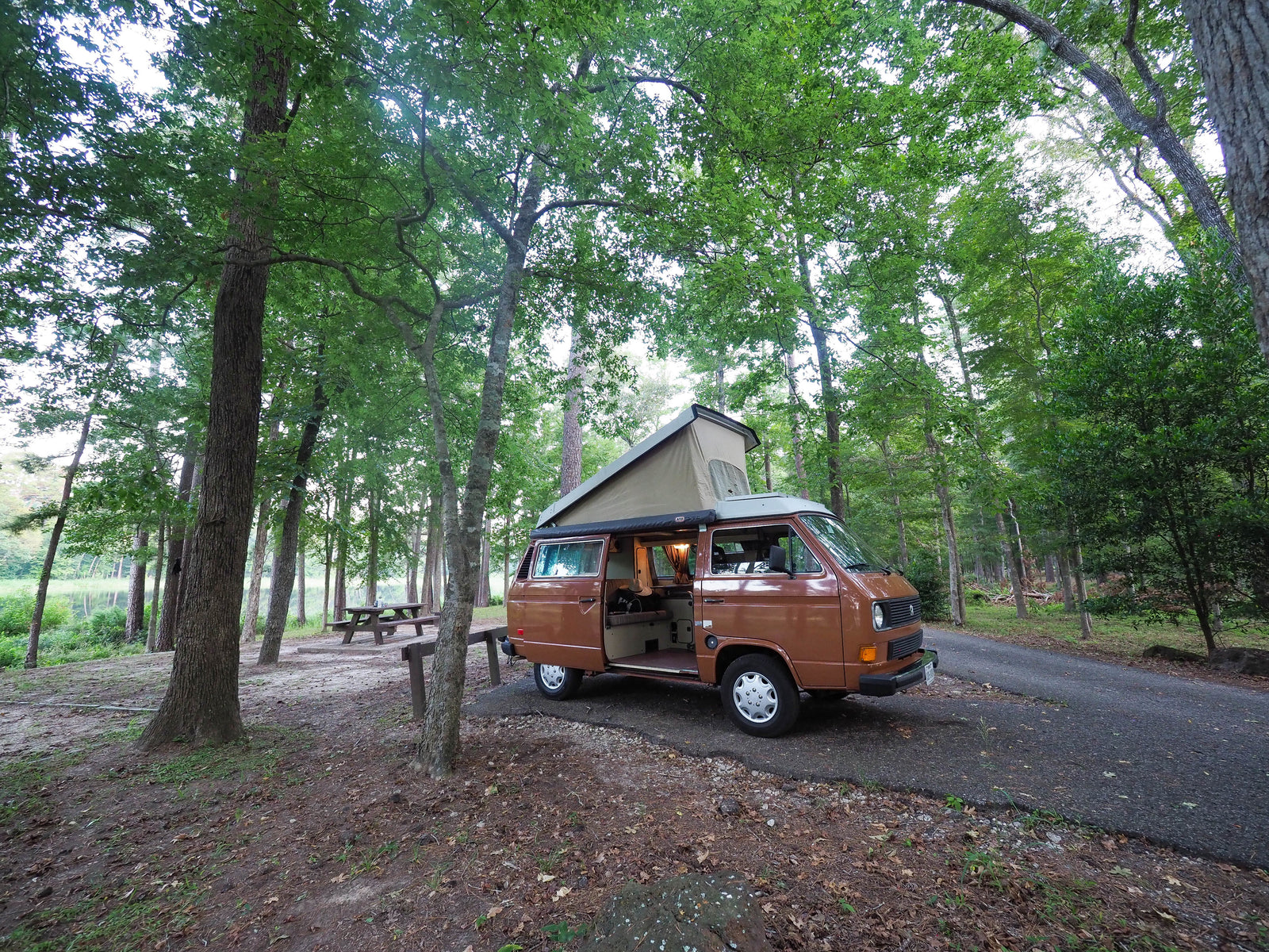 Experience camping! Brown RV rental (VW camper van) parked in a lush forest campsite by a lake. Interior light on.
