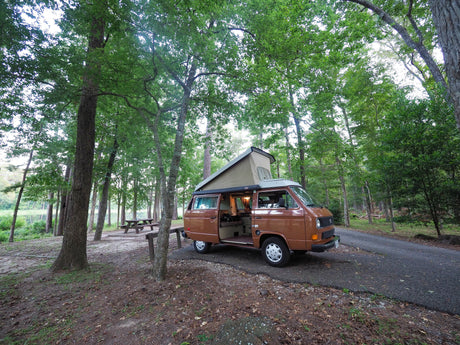 Experience camping! Brown RV rental (VW camper van) parked in a lush forest campsite by a lake. Interior light on.