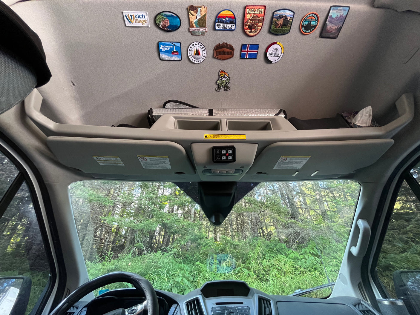 Inside an RV rental: dashboard, overhead storage, and travel patches from National Parks and destinations, with a forest view.