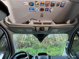 Inside an RV rental: dashboard, overhead storage, and travel patches from National Parks and destinations, with a forest view.