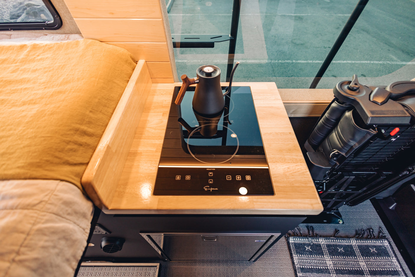 Modern RV rental interior: sleek induction cooktop with kettle, wooden countertop, cozy bed, and fridge.