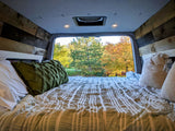 Cozy RV rental camper van interior. A bed with a green pillow and striped comforter offers a serene view of autumn trees.