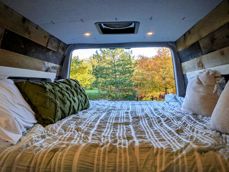 Cozy RV rental camper van interior. A bed with a green pillow and striped comforter offers a serene view of autumn trees.