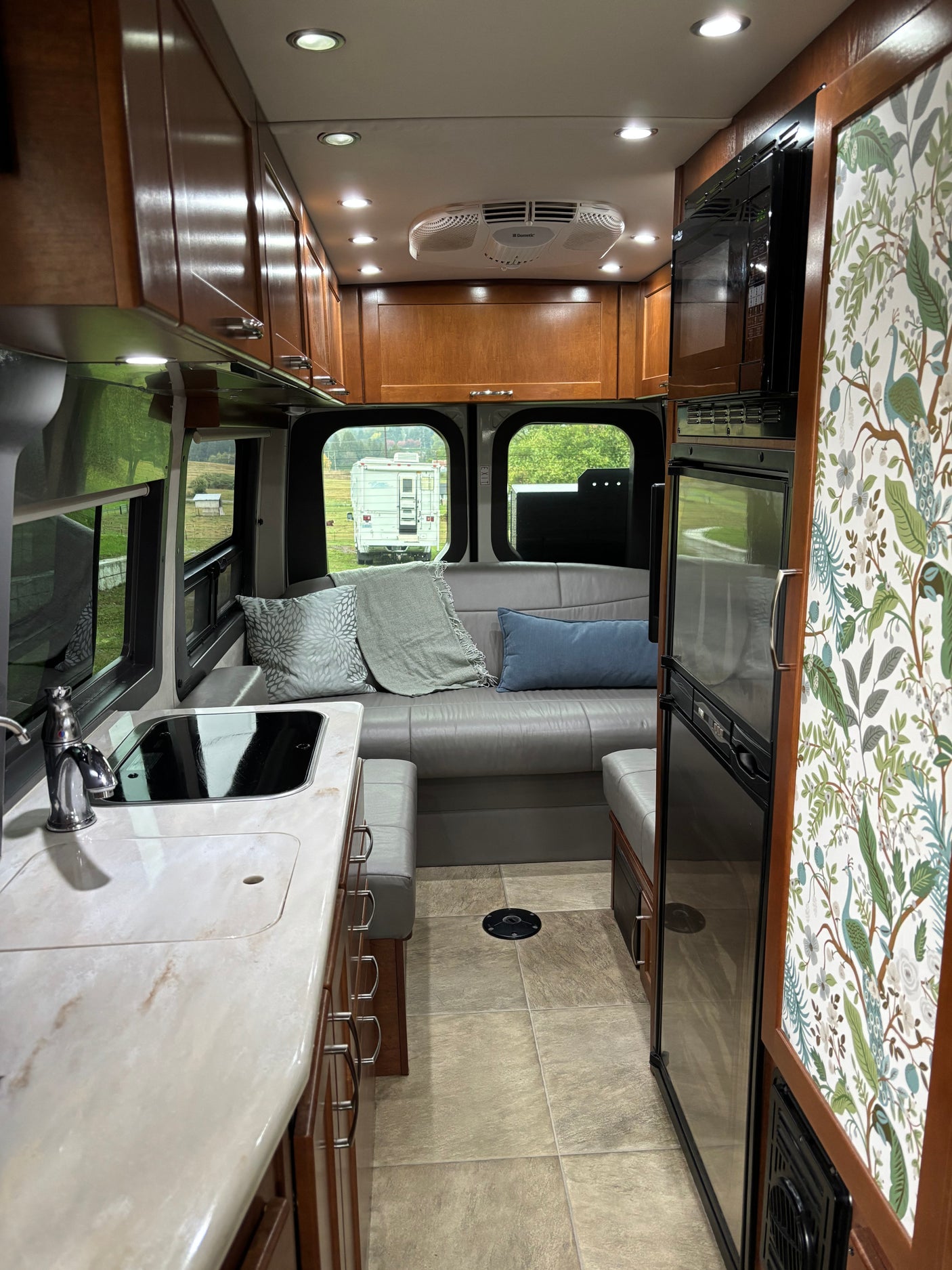 RV rental interior: a fully equipped kitchen with sink and stove, wood cabinetry, plush gray seating, and scenic views.