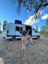 Relaxing van life with an RV rental. A girl stands while an adult reads by their white camper van in nature.