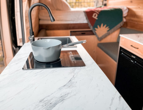 Inside this modern rv rental, a grey saucepan rests on an induction cooktop on a white marble-look counter. Sink and cozy bed visible.