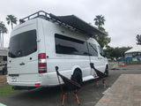 White Mercedes Sprinter RV rental van with an extended awning & camping chairs set up at a campsite.