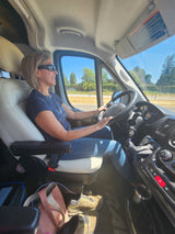 Woman in pilot seat of an RV rental van, smiling and driving. Bright interior shot during a sunny road trip.