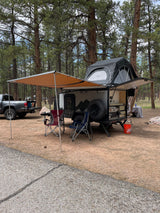 This RV rental overland trailer, featuring a rooftop tent and awning, is set up for camping in a peaceful pine forest.