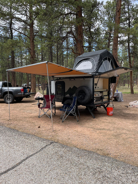 This RV rental overland trailer, featuring a rooftop tent and awning, is set up for camping in a peaceful pine forest.