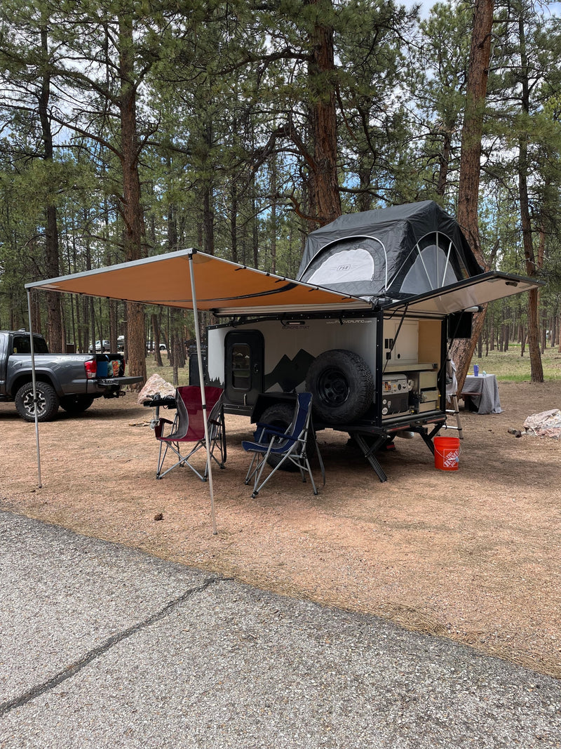 This RV rental overland trailer, featuring a rooftop tent and awning, is set up for camping in a peaceful pine forest. — photo 10