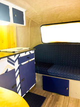 Inside a renovated RV rental: a modern blue & white kitchenette, cozy seating, and large window create a bright, compact space.