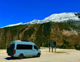 Light blue **rv rental** van at base of snowy mountains & forest. Sign for Illecillewaet Valley Trails. Scenic road trip adventure.