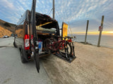 An RV rental, a converted van, is parked along a scenic ocean cliffside road at sunset, showcasing interior bed & bikes.