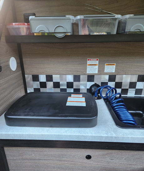 RV rental's compact kitchen features a covered stove, sink with blue hose, overhead storage, and checkerboard backsplash.