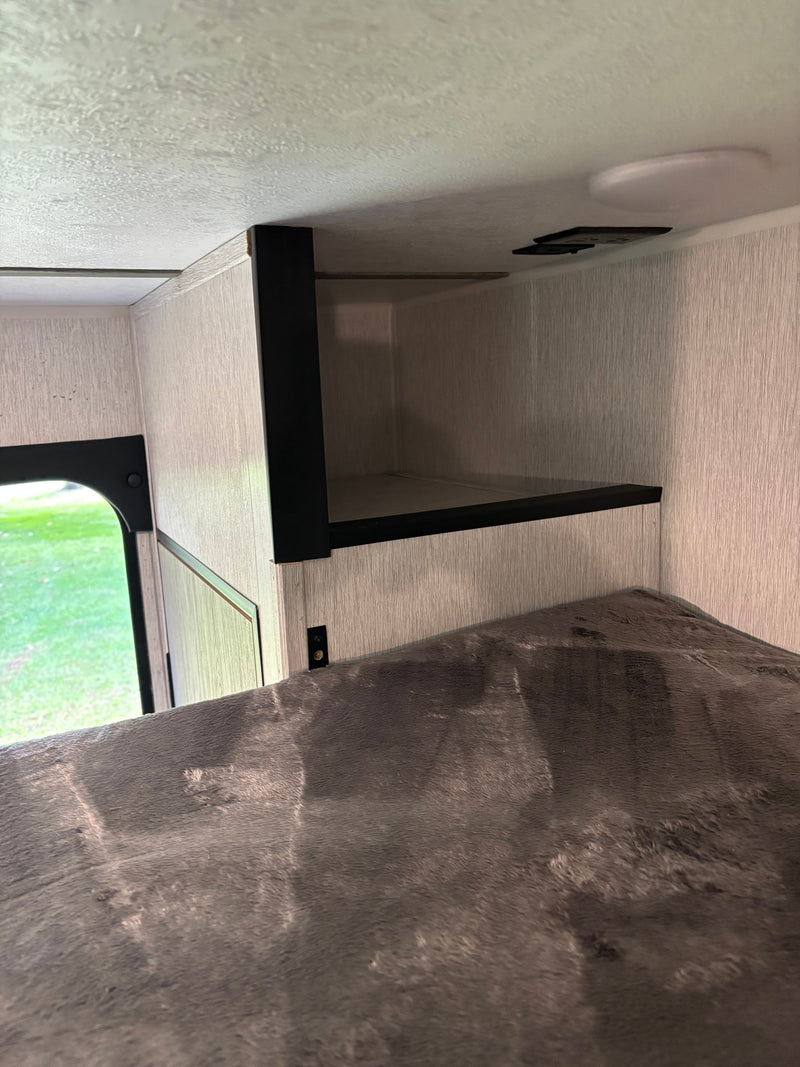 RV rental interior showing a cozy bunk bed with grey, textured bedding and an open black overhead storage shelf. — photo 13