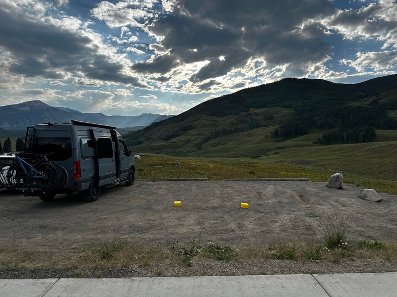 Grey RV rental campervan with bikes parked at a scenic mountain overlook under dramatic, cloud-filled skies over vast green hills. — photo 2