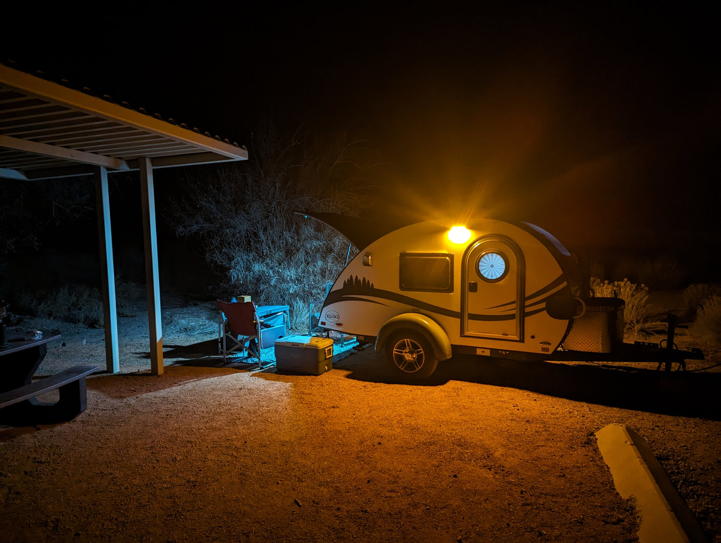 Nighttime RV rental campsite: a cozy teardrop trailer glows warmly next to a picnic table and cooler under a shelter.