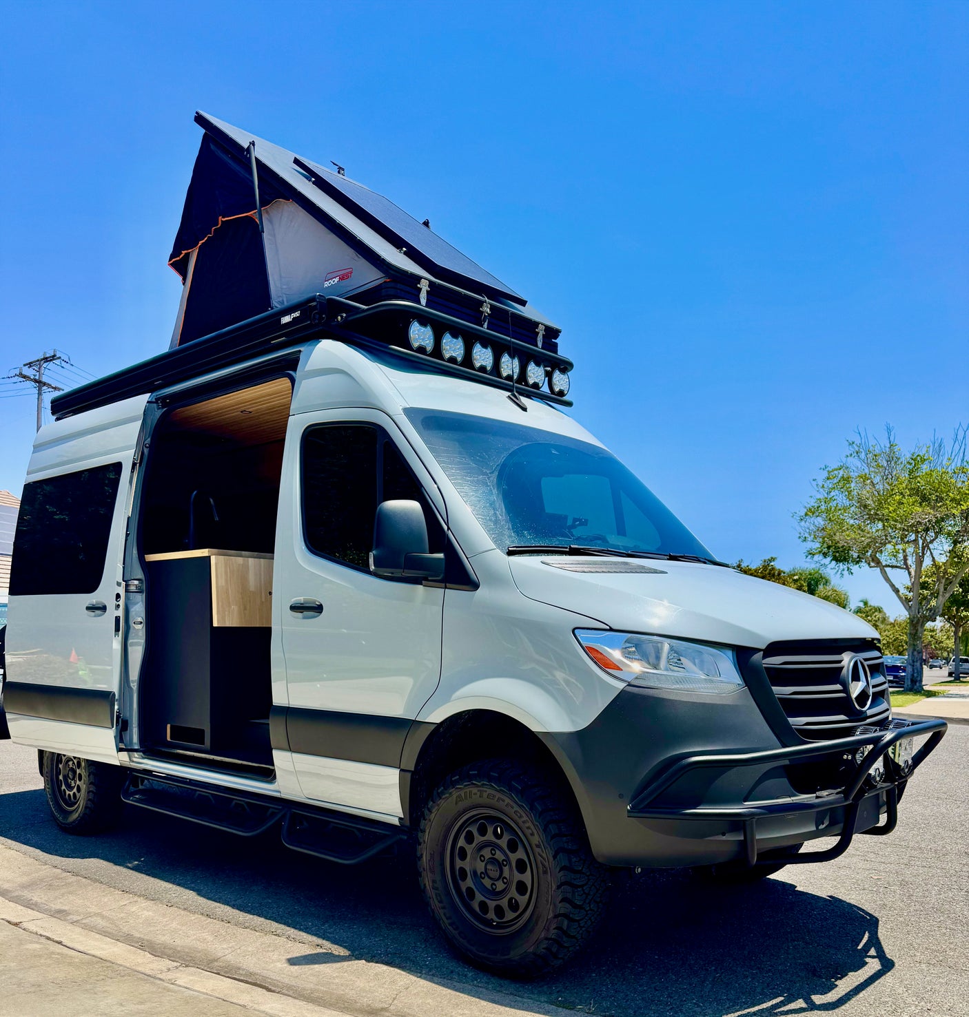 Mercedes-Benz Sprinter RV rental: A custom camper van with a pop-top, off-road tires, & lights, parked under a blue sky.