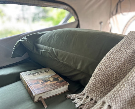 Inside an RV rental: a comfy bed with a green pillow, knitted blanket, and 'Wanderlust' book, hinting at adventure.