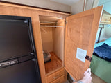 RV rental closet beside fridge, featuring a hanging rod, bin & drawers. Dinette conversion guide on door, with bed nearby.