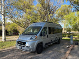 An RV rental, a silver Winnebago Travato, parked on a gravel campsite with lush trees and a blue sky overhead.