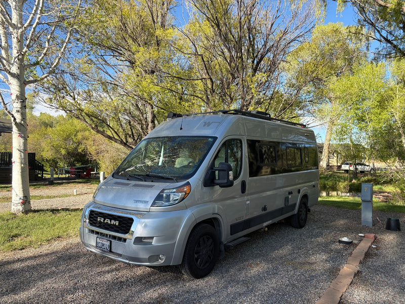 An RV rental, a silver Winnebago Travato, parked on a gravel campsite with lush trees and a blue sky overhead. — photo 4
