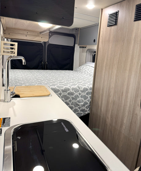 Interior of an rv rental: compact kitchen with cooktop, sink, and wooden cutting board. Beyond is a bed with gray patterned comforter.