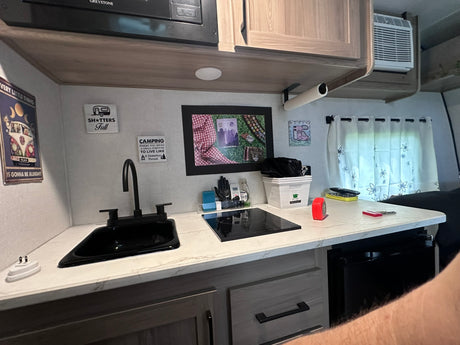  parked outdoors under a canopy with trees and a residential street in the background.|Coleman RV rental travel trailer parked on a sunny residential driveway. Notice its light exterior and leveling blocks.|Inside an RV rental: a bedroom with a black bed