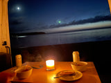 Cozy RV rental dinner by the water at night. Candlelight illuminates food, drinks, and a serene moonlit lake with a faint sky glow.