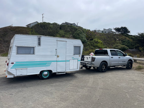 A silver pickup truck tows a white/teal travel trailer on dirt. Houses are visible on the green hill behind. Imagine an RV rental road trip!