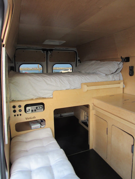 Cozy wood-paneled RV rental interior with a raised bed, control panel, seating, and a scenic view out the back windows.