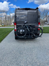 Ram ProMaster RV rental van with mountain bike on hitch rack, parked on a residential driveway.