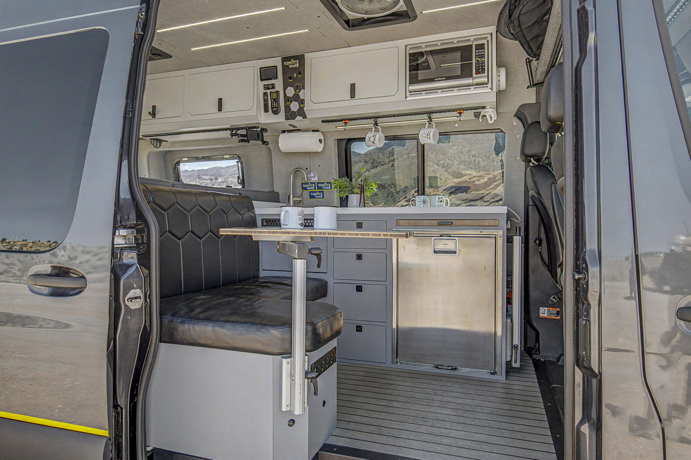 Inside a modern rv rental camper van, a sleek kitchen, dining area, and scenic mountain views are seen from the open side door.