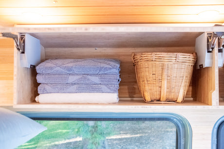 Cozy RV rental interior: overhead storage with neatly folded blue blankets and a wicker basket. Window view of greenery.