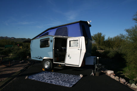 Pop-up RV rental: Light blue camper with open door at a scenic desert campsite, ready for your outdoor adventure.