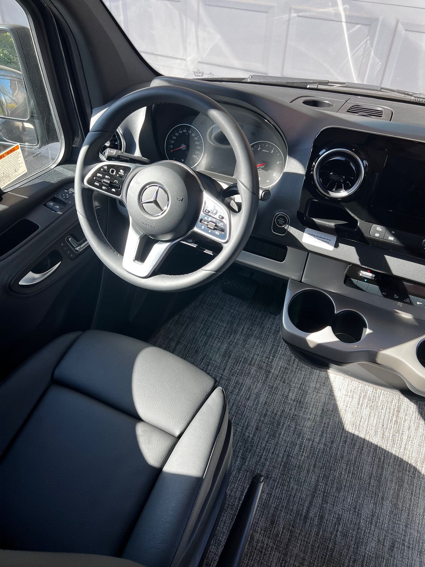Driver's view of a Mercedes-Benz van interior for rv rental, with steering wheel, dash, cup holders, and gray floor.