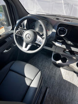 Driver's view of a Mercedes-Benz van interior for rv rental, with steering wheel, dash, cup holders, and gray floor.