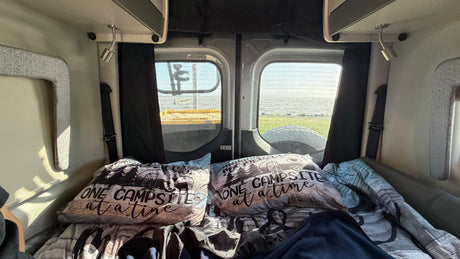 Cozy RV rental bed, 'ONE CAMPSITE at a time' pillows, with a scenic view of water and grass through the rear windows.
