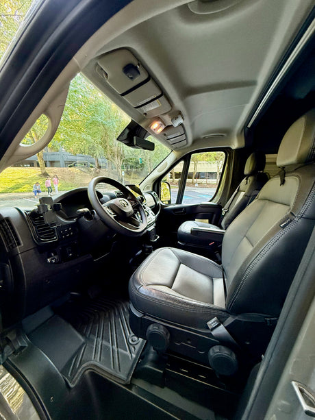 Inside this modern RV rental van, the driver's and passenger seats, dashboard, and a view of trees and people outside are visible.
