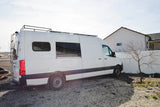 A white Sprinter camper van with a roof rack and ladder, parked on gravel next to a house. Ideal for an RV rental adventure.